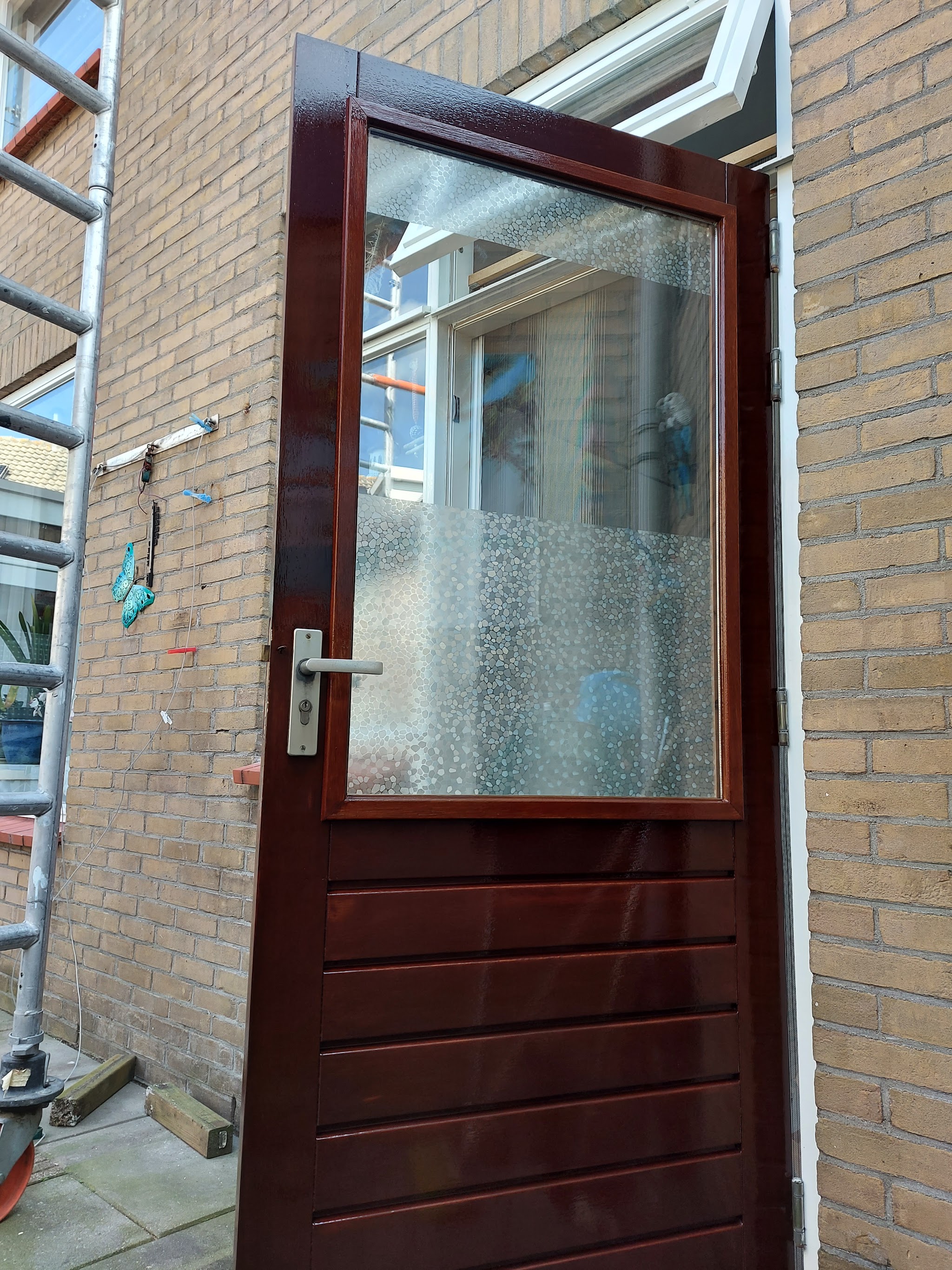 Pas gelakte houten achterdeur in donkerrood met glas, naast een bakstenen muur