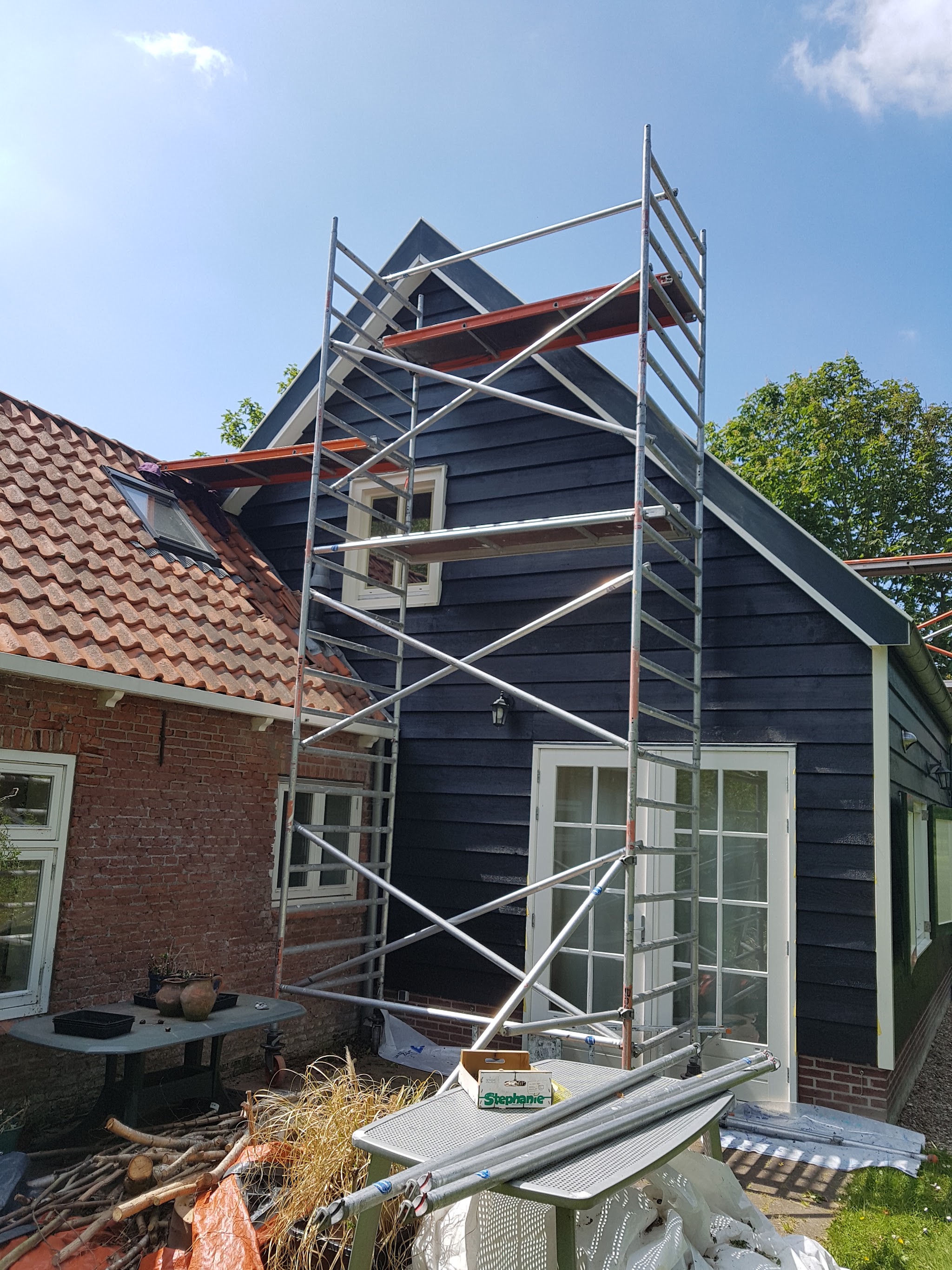 Donker geschilderde houten topgevel met witte kozijnen, met een rolsteiger ervoor